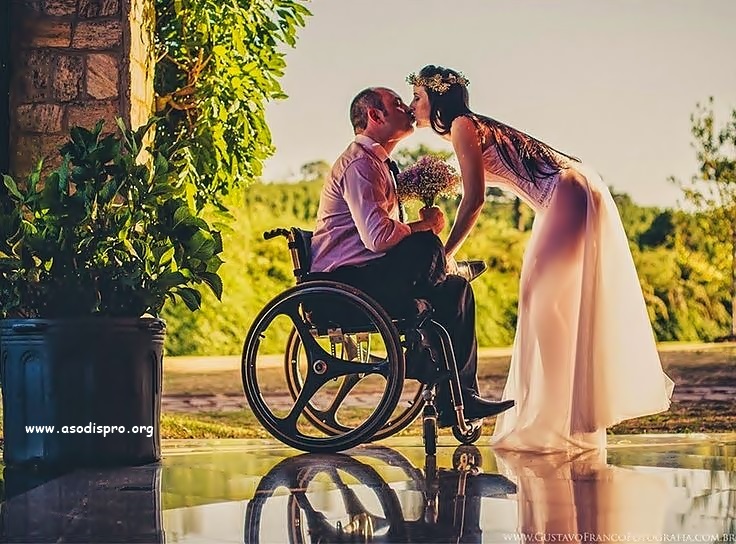 Una pareja de novios se besa, él en silla de ruedas sostiene el ramo de flores, mientras ella agachada resalta un vestido casi transparente por la luz del sol de una tarde. 