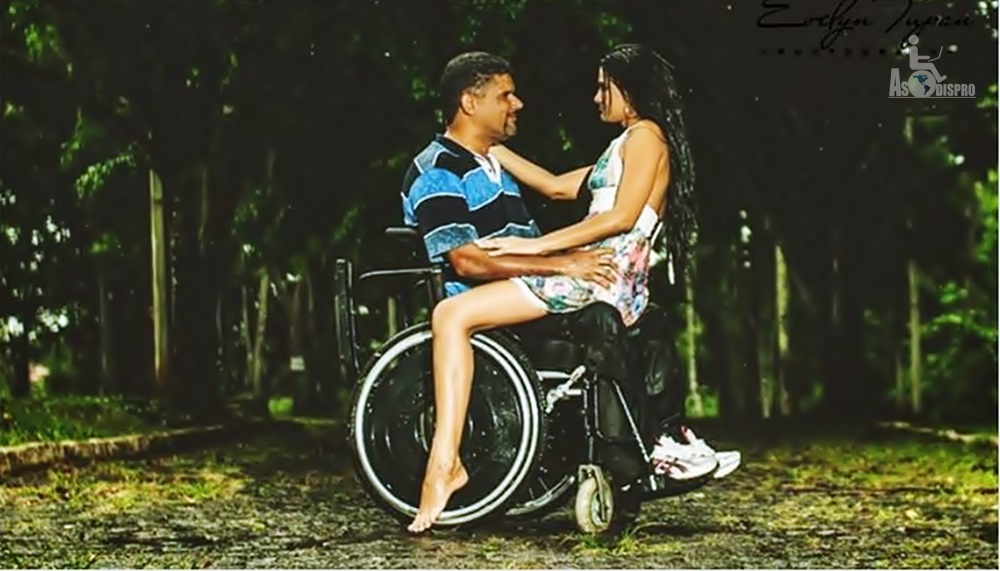En un bosque, un hombre en silla de ruedas carga sobre sus piernas a una mujer.