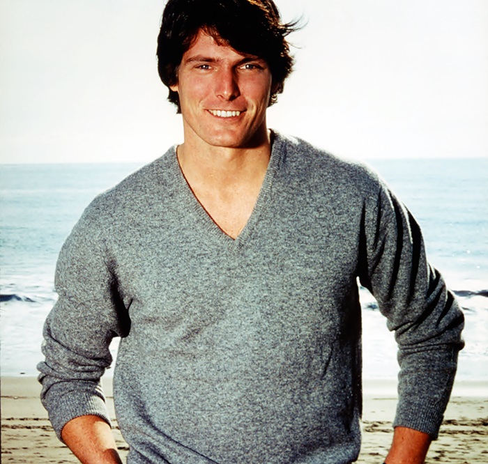 Christopher Reeve posa con las manos en la cintura, esto vestido con un sudadero y teniendo de fondo una playa.