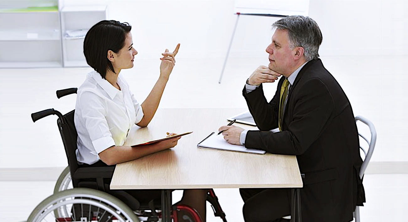 Una mujer en silla de ruedas está de frente a un ejecutivo en un escritorio, él pone atención y ella habla.