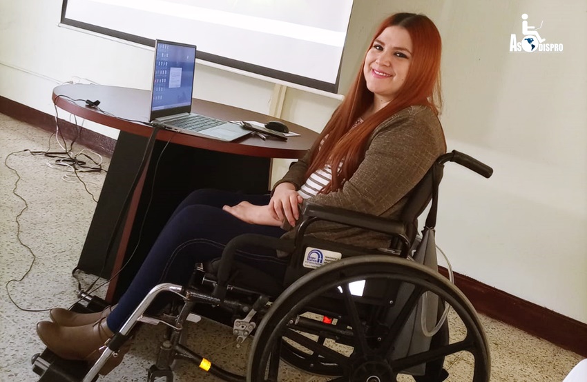 En su silla de ruedas y frente a una mesa de presentación con una computadora y una pantalla al fondo, Lucrecia sonríe previo a iiniciar la reunión.