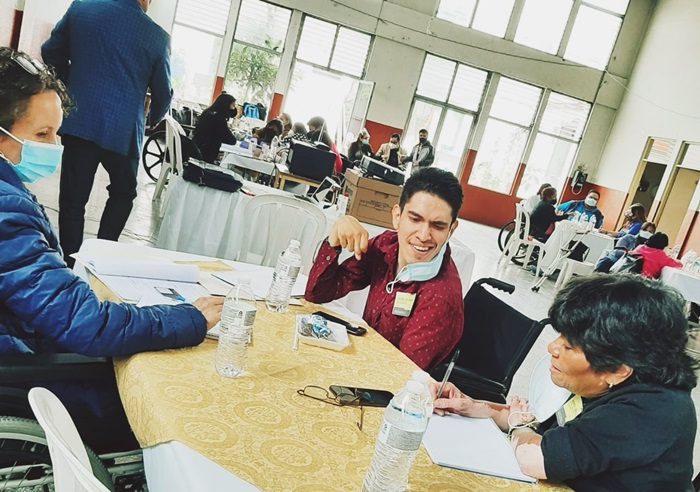 En una mesa, 3 personas con discapacidad interactuan mientras una escribe. Al fondo se ve más mesas con participantes. 