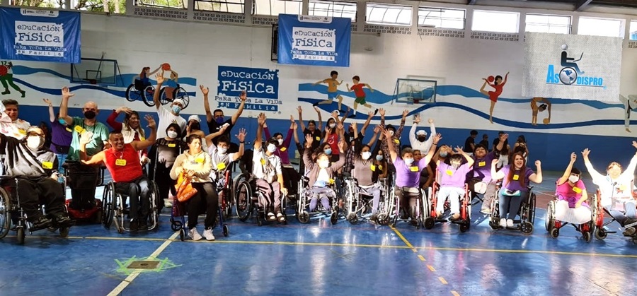 En un gimnasio unas 50 personas con discapacidad levantan las manos alegremente, esto en un evento de ASODISPRO.