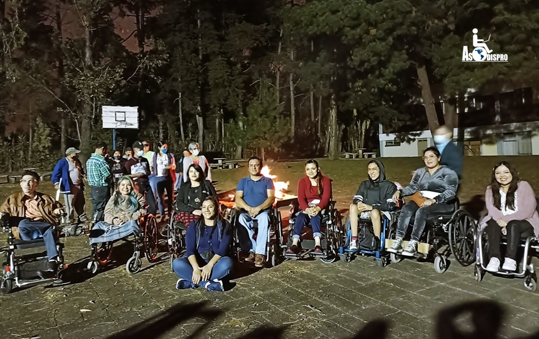 Fotografía de noche en un campo con árboles, con personas y una fogata al fondo, se ven 8 personas en sus sillas de ruedas, y la coordinadora Alba Velásquez, sentada al frente en el suelo.
