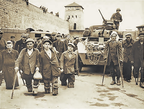 En la fotografía en blanco y negro, se observan varios prisioneros de la segunda guerra mundial, caminan frente a un tanque mientras son observados por un soldado que sobresale del tanque.