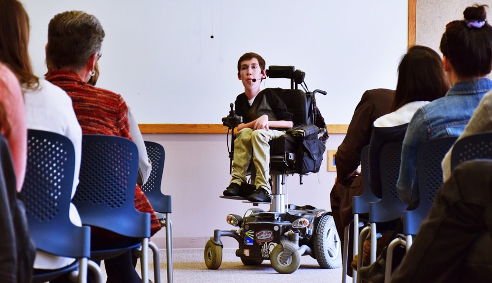 Shane Burcaw está con un micrófono inalámbrico, en su silla de ruedas en un salón de clases frente a un público que le observa con atención.