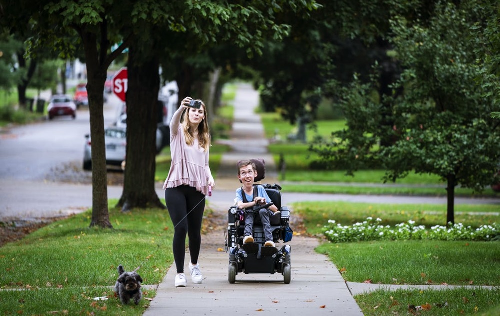 En una acera de un área residencial, se observa a Hannah Aylward tomando un selfie mientras camina a la par de Shane en su silla de ruedas y un pequeño perro. 