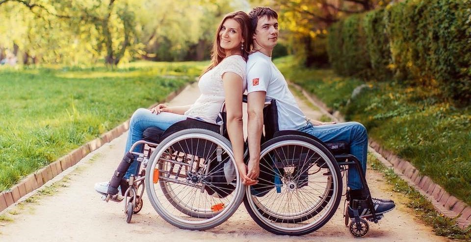 Una pareja de jóvenes en silla de ruedas, mujer y hombre, están de espaldas uno contra otro, sonríen mientras ven a la cámara tomados de la mano en una vereda. 