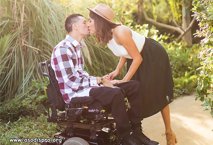 Un hombre en silla de ruedas eléctrica besa a una chica, que se agacha un tanto para besarle.