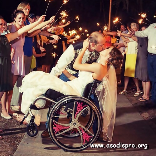 Vestidos para casarse, el novio sostiene a la novia un tanto recostada para atrás con su silla de ruedas mientras se besan y al fondo los invitados sostienen luces de pólvora. 