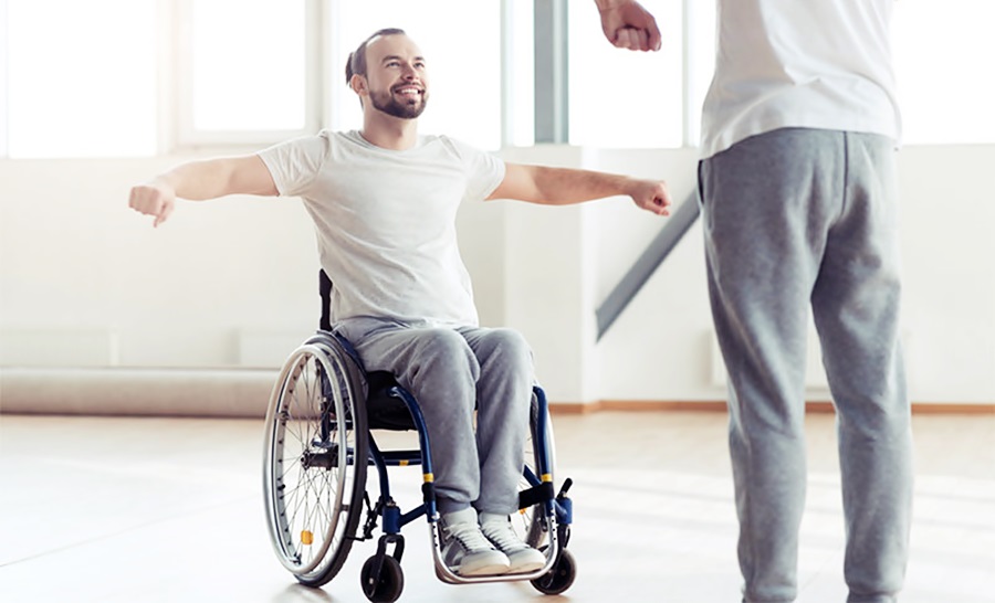 Un hombre en silla de ruedas abre sus brazos, haciendo gimnasia frente a otra persona de pie.