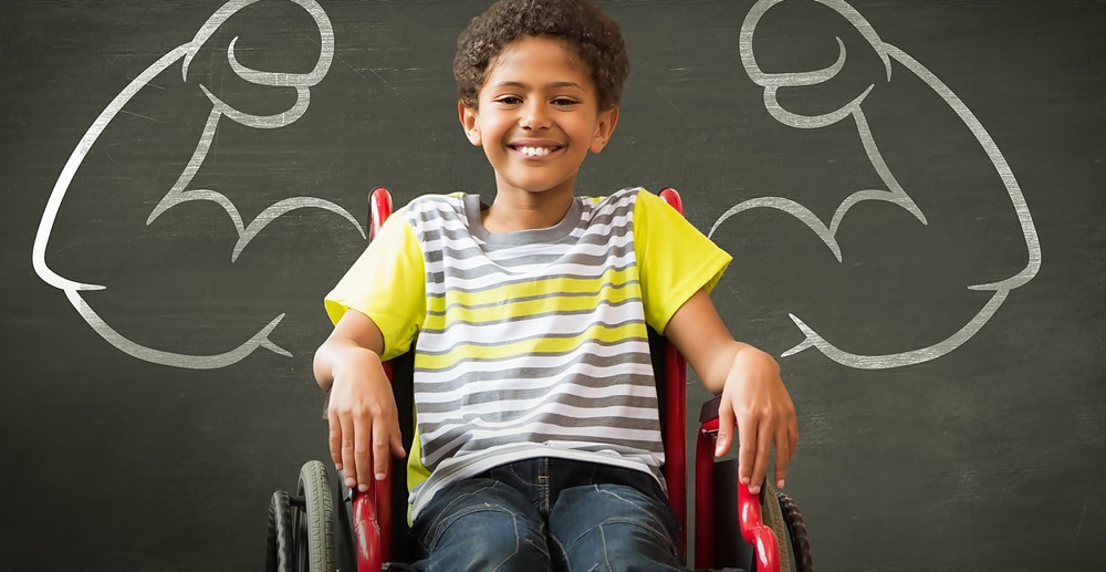 Un niño sonriente con discapacidad física está sentado en una silla de ruedas frente a un pizarrón que tiene dibujado dos brazos musculosos y pareciera que salieran del infante. 