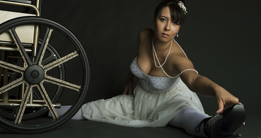 una joven mujer está en el suelo, vestida con un traje de bailarina, estira sus piernas. A la par se encuentra una silla de ruedas.
