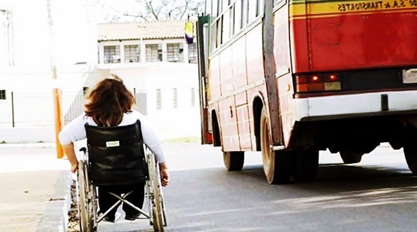 Una mujer en silla de ruedas va por una calle y a su par, pasa un bus colectivo. 