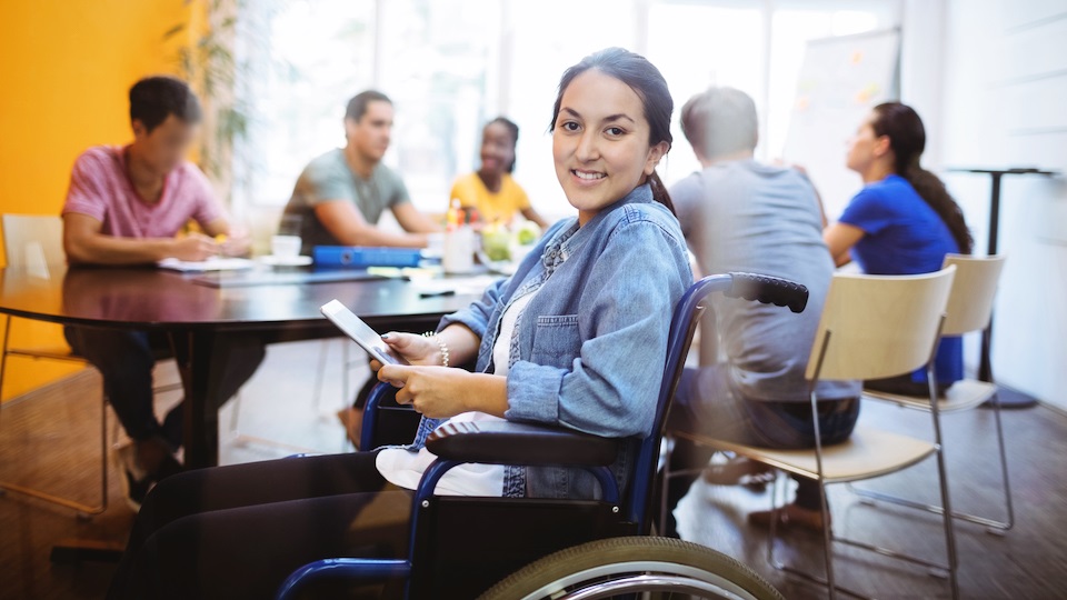 Una chica sonriente en silla de ruedas sonríe mientras atrás algunas personas trabajan en una mesa.