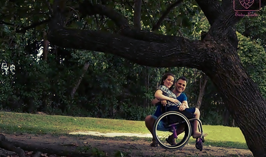 En un claro de un bosque, una mujer en silla de ruedas sonríe a cámara junto a su pareja quien está a su lado. 