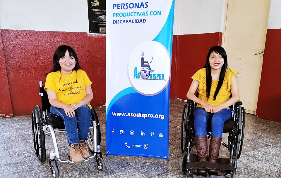 Las 2 chicas posan sonriendo en sus sillas de ruedas y con un cartel con el logo de Asodispro atrás de ellas.