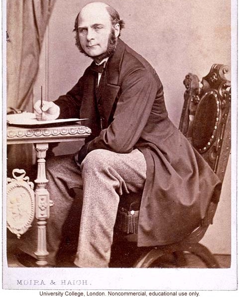 Sentado en la silla de un escritorio, Galton escribe en un folder mientras ve a la cámara, vistiendo de corbata y gabardina. 