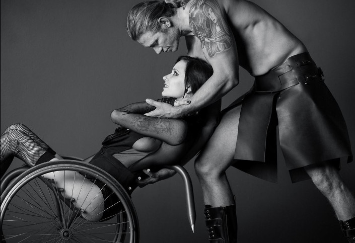 Una mujer en lencería negra, se recuesta de espaldas con su silla de ruedas mientras un hombre la sostiene con el torso descubierto, de pelo largo, y con una falda (al parecer de cuero).