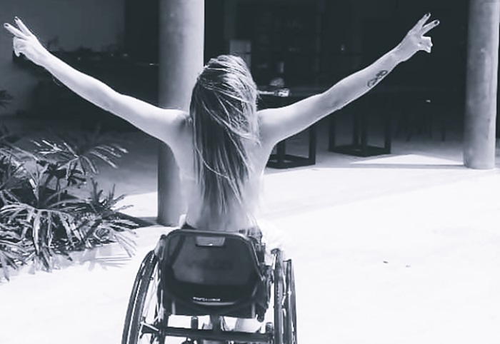 En una fotografía en blanco y negro, una mujer sin ropa, de pelo largo y de espaldas en una silla de ruedas. 