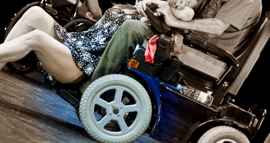 De medio cuerpo se ve a una mujer con vestido muy corto recostada en el regazo de un hombre en silla de ruedas eléctrica 