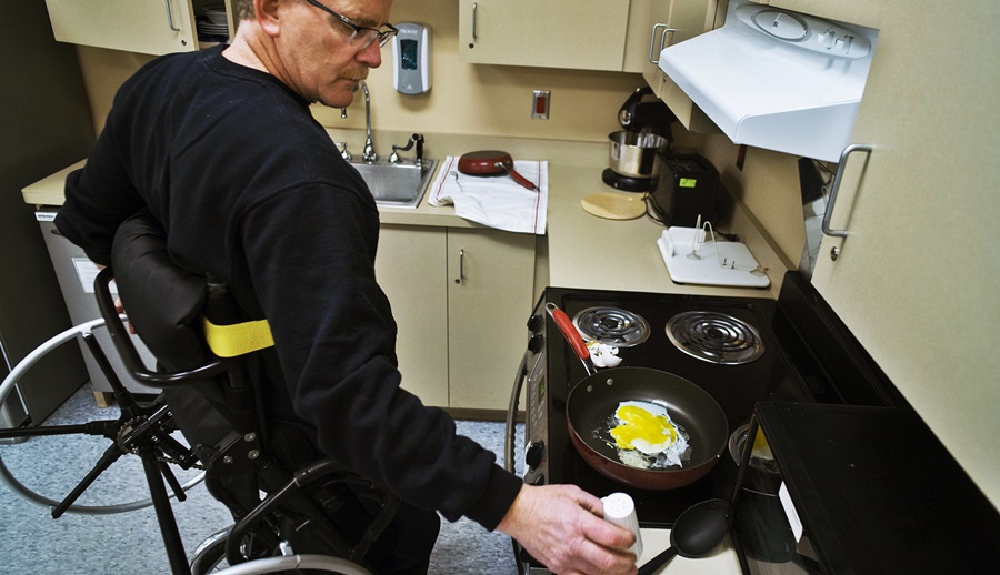 Utilizando una silla de ruedas que lo pone de pie, un hombre prepara unos huevos en una cocina 