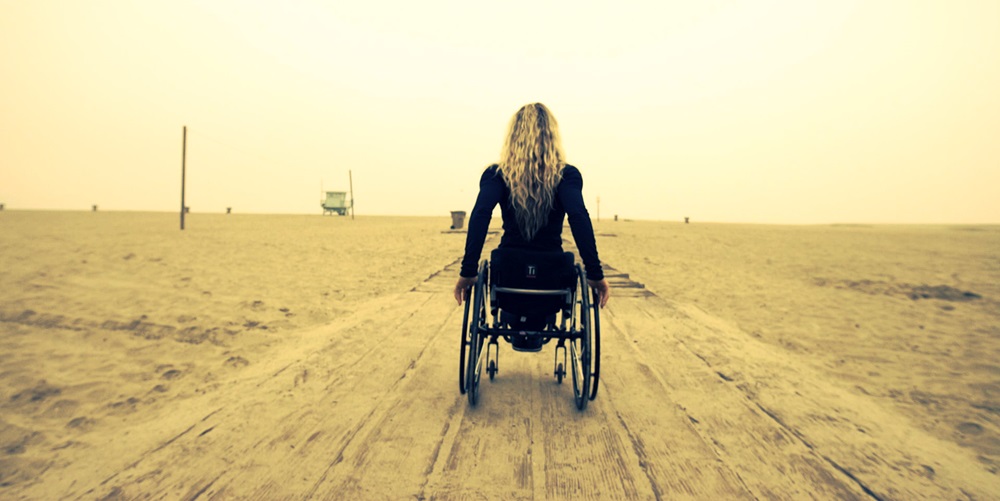 De espaldas se ve a Adams quién va en su silla de ruedas en una playa 