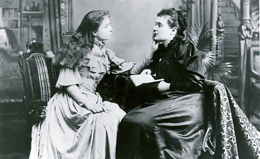 Sentadas en lo que parece una sala, las jóvenes Helen Keller y Anne Sullivan, esta última sostiene un libros que al parecer le lee a Helen, mientras esta le toca el rostro con una de sus manos.