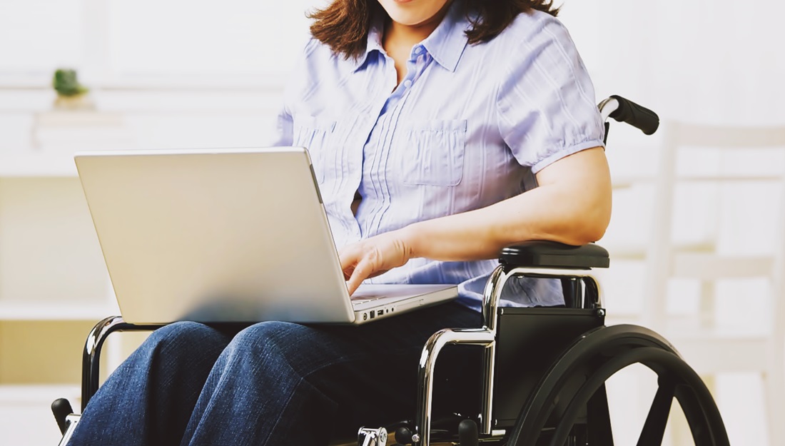 Fotografía del cuerpo de una mujer sentada en su silla de ruedas, escribiendo en una computadora que está en sus piernas 