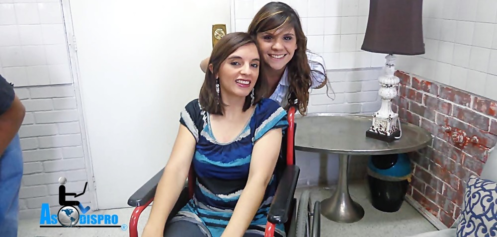 Sentada en su silla de ruedas, Mónica sonríe a cámara con la entrevistadora tras ella y sonriendo igualmente a cámara