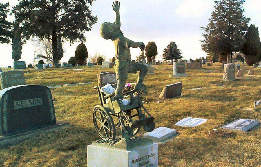 En un cementerio con varias lapidas a ras de suelo, destaca la estatuas de un niño parado en un pie sobre una silla de ruedas, alzando una de sus manos cómo si esperara ser halado hacia arriba, su rostro está viendo al cielo. 