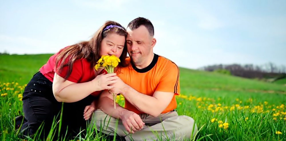 Una niña y un adulto con Síndrome de Down huelen unas flores en una pradera