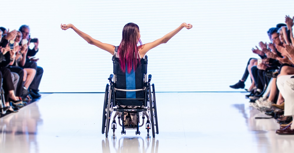 Una mujer con tetraplejía levanta las manos, en su silla de ruedas, mientras un público a los costados aplaude. Al parecer es una pasarela de modas, la fotografía se tomó con la chica de espalas. 