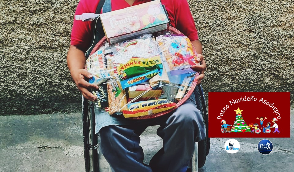 Una persona en silla de ruedas sostiene una canasta navideña con varios productos. La fotografía está cortada para que no se vea la cara del beneficiado. 