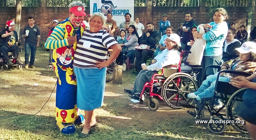 Doña Marce posa cómicamente junto a un payaso, mientras entre el público su hija se ríe de la escena entre el público de un Paseo Navideño Asodispro.