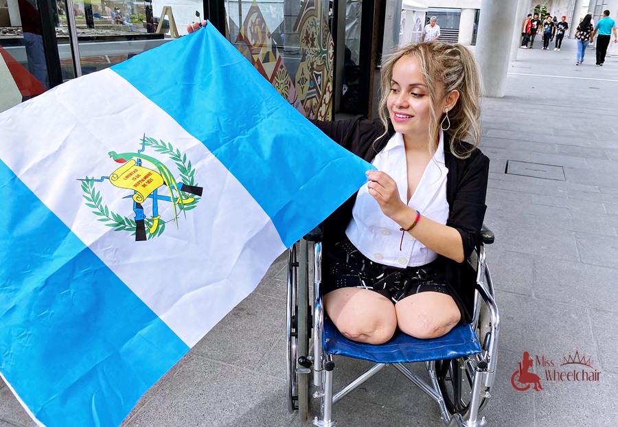 En una calle, María está en su silla de ruedas, de blusa blanca, saco negro y short negros, sonríe mientras con sus manos sostiene la bandera de Guatemala.  