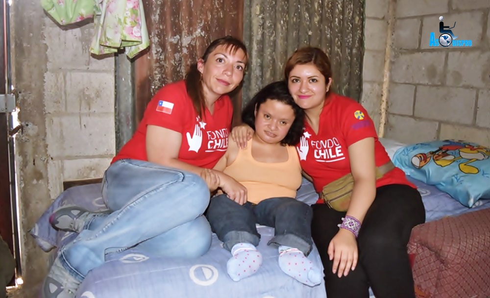 En una cama, dos delegadas chilenas con playeras del programa, posan junto  Gaby, persona con discapacidad del área social de Asodispro