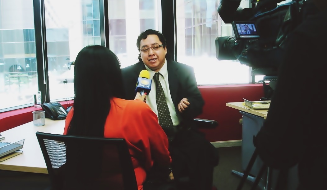 En una oficina, Byron Pernilla, en su silla de ruedas eléctrica y de saco y corbata, es entrevistado por una reportera con el micrófono de Noticieros Televisa México