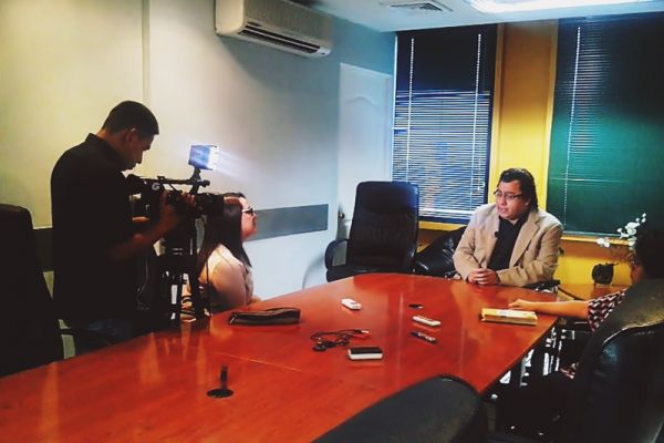 En una sala de juntas, Byron Pernilla es entrevistado por una periodista y un camarógrafo.