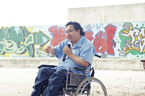 Micrófono en mano y en su silla de ruedas, Byron Pernilla desarrolla una charla en un lugar al aire libre, al fondo se observa una pared con grafitis. 