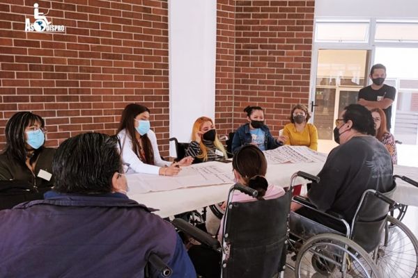 En una mesa de trabajo, y en sus sillas de ruedas, las personas con discapacidad socias de ASODISPRO trabajan durante el taller de réplica.