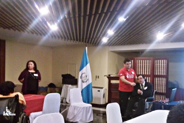 En un salón de un hotel, con la bandera de Guatemala, Byron Pernilla habla por medio de un micrófono que sostiene un asistente.