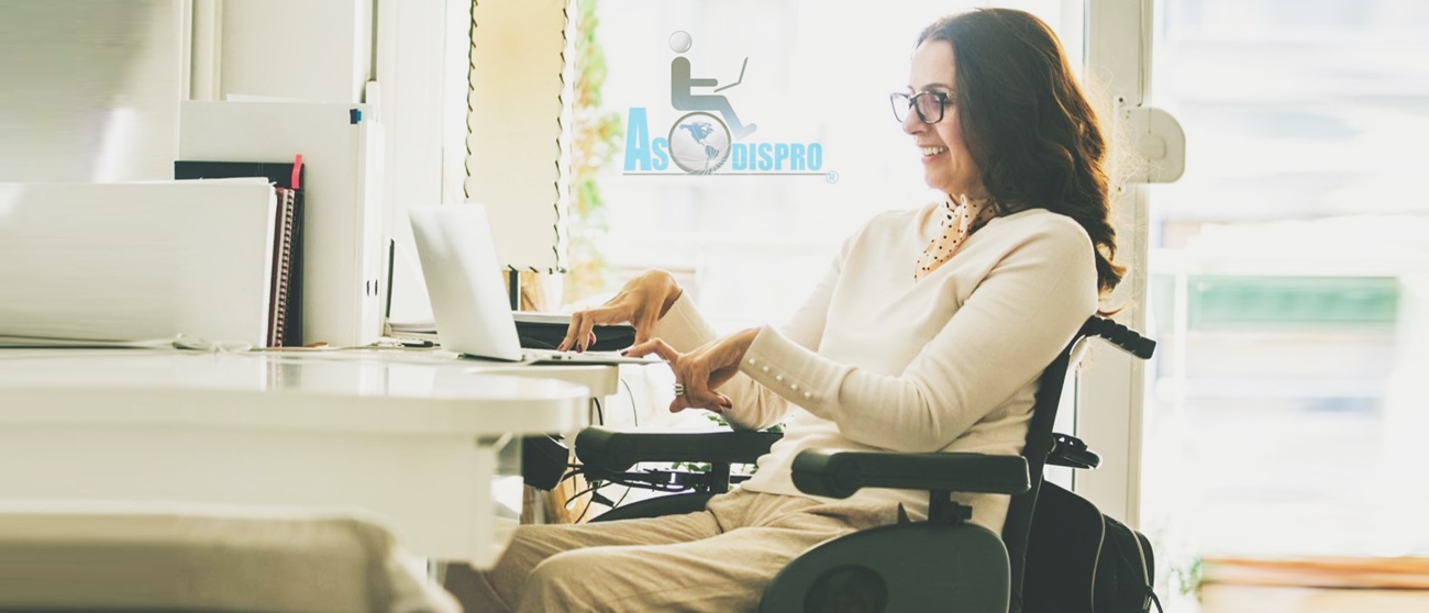 En una oficina, una chica en silla de ruedas eléctrica, sonríe mientras trabaja en una computadora en un escritorio, al fondo el logo de ASODISPRO 