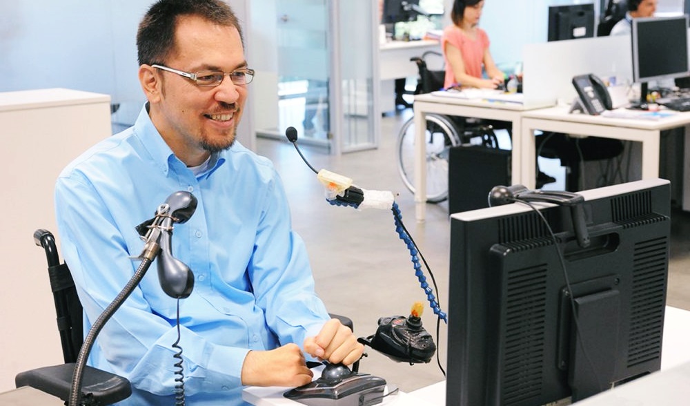 Un hombre tetraplejico trabaja en un escritorio adaptado, al fondo se observa a otra trabajadora en silla de ruedas