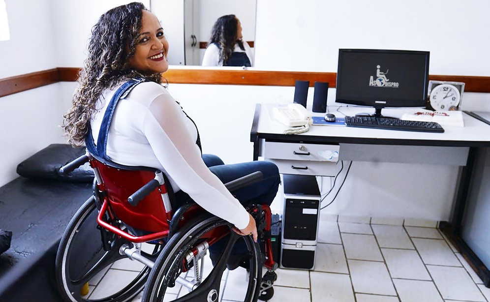 Una joven mujer en silla de ruedas, voltea y sonrie a cámara frente a un escritorio con computadora 