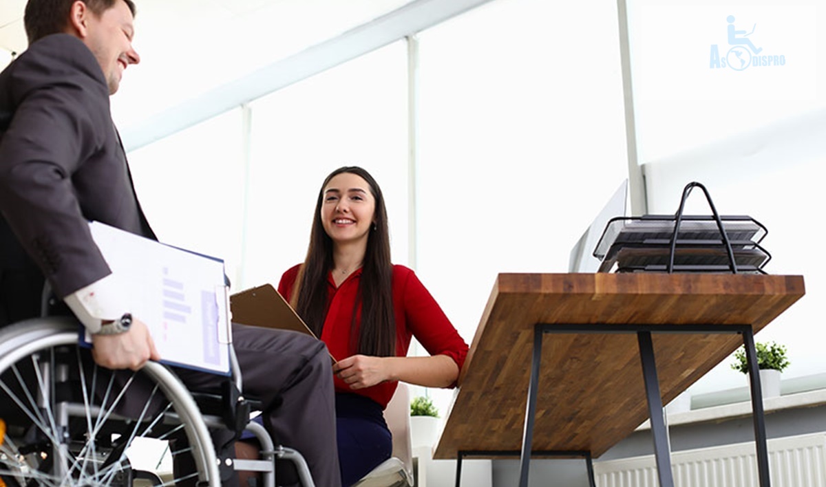 Una chica ejecutiva sentada sonríe a un joven en silla de ruedas que llega portando documentos