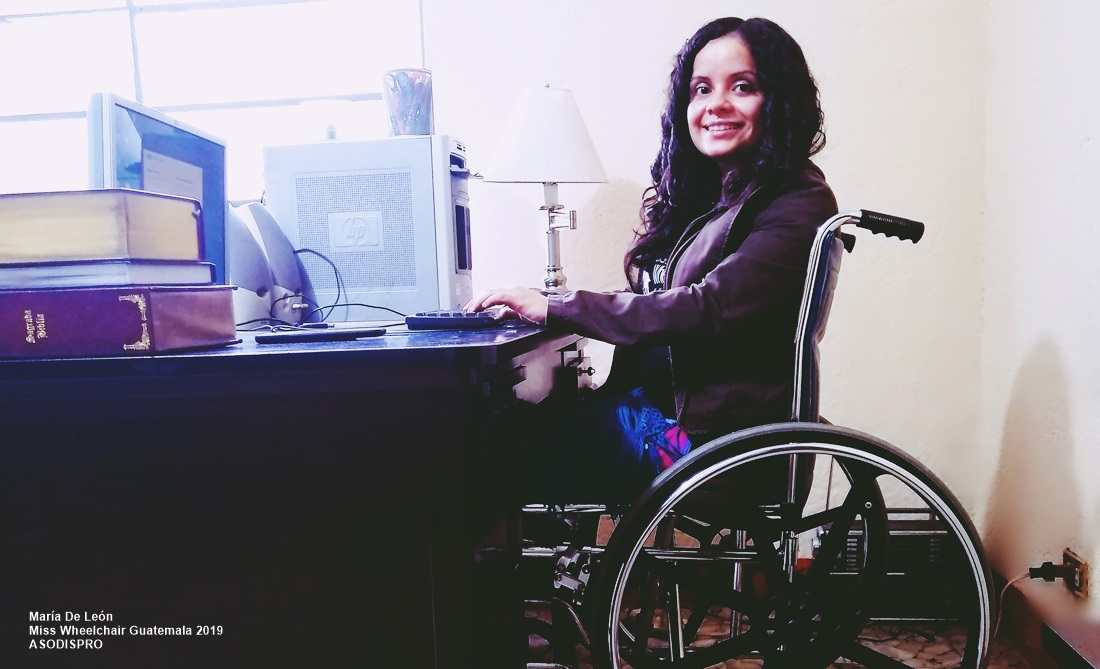 María De León, sentada en su silla de ruedas, está en un escritorio frente a una computadora y sonríe a cámara.
