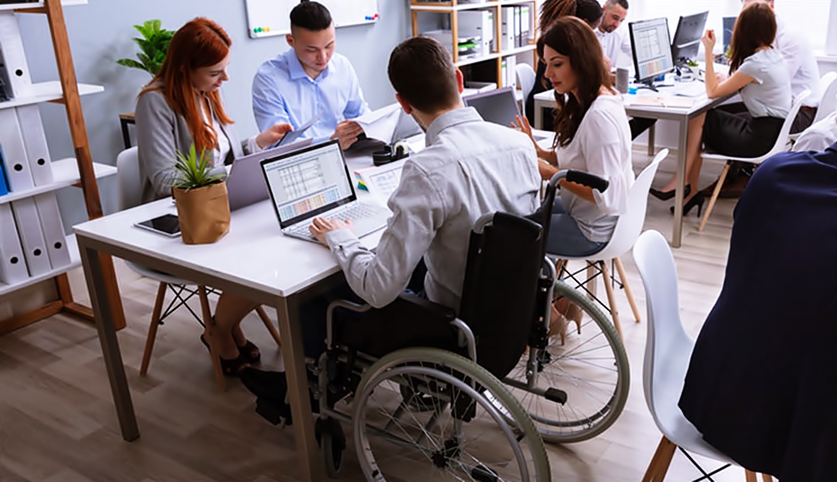 En una sala de trabajo, varios ejecutivos trabajan en sus computadoras, uno de ellos usuario de silla de ruedas.