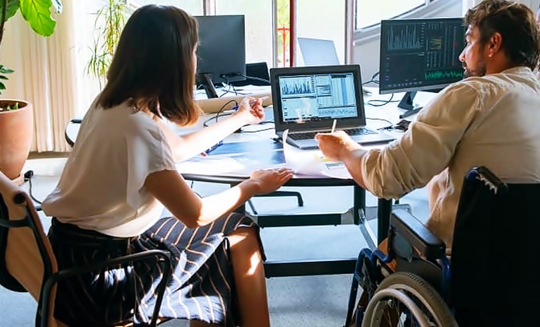 Un hombre y una mujer, él en silla de ruedas y ella en una silla, conversan frente a una mesa de trabajo con una computadora.