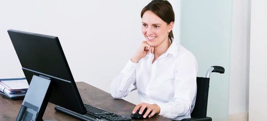 Frente a una computadora en un escritorio, una señorita en silla de ruedas sonríe al trabajar